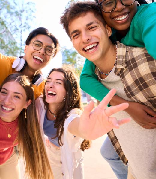 Vertical multiracial group of teenagers taking a selfie looking front camera laughing and having fun piggybacking together outdoor. Young students enjoying their free time with friends in street city
