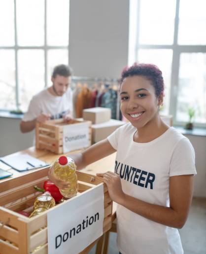 Help, support. Young laughing happy girl volunteer standing near a filled donation box with a bottle in his hand, a man packing boxes.